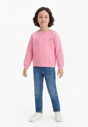 Barn med skulderlangt hår iført en rosa genser med hjertedetalj, blå jeans og hvite joggesko, stående foran hvit bakgrunn.