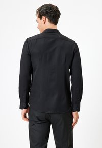 Camicia nera a maniche lunghe con un tessuto testurizzato, polsini con bottoni e colletto strutturato, mostrata dal retro. Design liscio e aderente.