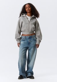 Γκρι κοντό φούτερ με φερμουάρ και δύο μπροστινές τσέπες, συνδυασμένο με άνετο, ανοιχτό μπλε τζιν παντελόνι. Το μοντέλο φοράει μαύρα παπούτσια.