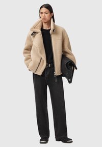 Beżowa kurtka z sztucznego futra z wysokim kołnierzem, zamek błyskawiczny i duże kieszenie, zestawiona z czarnymi spodniami typu wide-leg i czarnym obuwiem.