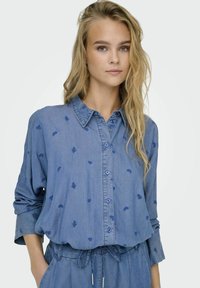 Camicia in denim blu con maniche lunghe, chiusura a bottoni e motivo floreale ricamato. Tessuto morbido, vestibilità ampia e design del collo classico.