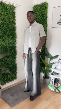 Giovane uomo che sta in piedi all'interno, indossando una camicia a maniche corte con un motivo, una maglietta bianca, jeans larghi sbiaditi, con le mani in tasca, vicino a piante verdi.
