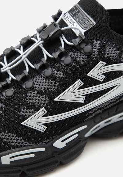 Μαύρο και γκρι πλεκτό αθλητικό παπούτσι με λευκά βέλη στο πλάι, μαύρα κορδόνια με κλιπ, και μαύρη λαστιχένια σόλα με λευκές λεπτομέρειες.
