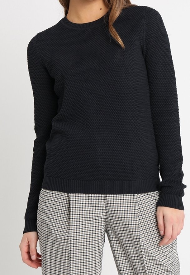Femme portant un pull noir texturé à manches longues, associé à un pantalon taille haute à carreaux noir et blanc.