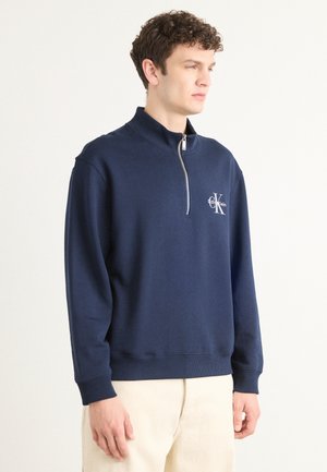 Navyblauwe pullover sweatshirt met een kwart rits, geribbelde mouwen en een klein wit logo op de borst. Zachte stof en een relaxte pasvorm.