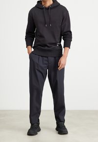 Muž ve černé mikině s kapucí s logem Calvin Klein, tmavě modrých plisovaných kalhotách a černých turistických botách stojící na světlé podlaze.