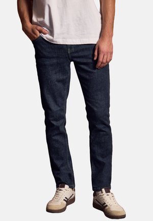 Jean en denim bleu foncé avec une coupe slim, présentant un design classique et une légère texture. À assortir avec des baskets beiges et marron.
