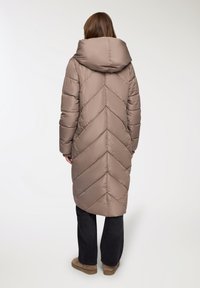 Długi, jasnobrązowy puchowy płaszcz z wzorem w zygzak, dużym kapturem i smukłą sylwetką. Noszony z ciemnymi spodniami i beżowym obuwiem.