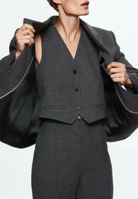 Personne ajustant un blazer cintré gris foncé par-dessus un gilet boutonné assorti et un pantalon, sur un fond blanc uni.