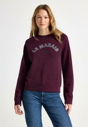 Felpa bordeaux con "LE MARAIS PARIS" ricamato in bianco. Presenta un collo rotondo e polsini a costine, abbinata a jeans in denim blu.