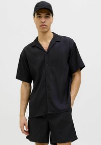Ensemble composé d'une chemise noire à manches courtes et d'un short, avec une texture côtelée verticale, comprenant un col et une fermeture à boutons, porté avec une casquette noire.