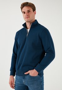 Sudadera azul marino con cremallera, hecha de tela suave, con un cuello alto y puños acanalados, combinada con jeans azules.
