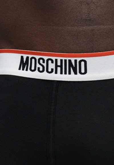 Ζώνη μαύρου εσωρούχου με λευκή ταινία που διαθέτει κόκκινη πάνω ρίγα και έντονο μαύρο λογότυπο "MOSCHINO" στο μπροστινό μέρος.