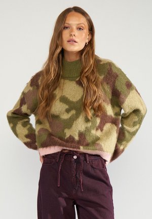 Vrouw met lang haar die een groen en bruine camouflage trui draagt en donkerviolette hoge taille broek, staand met handen in de zij.
