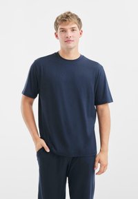 T-shirt a maniche corte blu navy in un tessuto liscio. Collo rotondo, vestibilità rilassata, con una leggera lucentezza. Il modello ha le mani in tasca e indossa pantaloni abbinati.