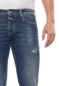Jeans en denim bleu foncé avec accents déchirés, dotés d'une fermeture à bouton, d'un design à cinq poches et d'un léger dégradé sur les jambes.