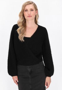 Blonde Frau mit welligem Haar trägt einen schwarzen, flauschigen Wickelpullover über einem schwarzen Oberteil und einem hoch taillierten, dunklen Jeansrock und steht vor einem weißen Hintergrund.