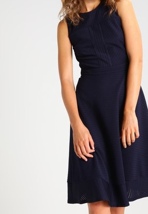 Granatowa sukienka bez rękawów z teksturowanej tkaniny, dopasowanym gorsetem i rozkloszowanym dołem. Charakterystyczne detale to panele i subtelne perforacje.