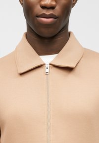 Κοντινό πλάνο ενός ατόμου που φοράει μπεζ ζακέτα με φερμουάρ και γιακά πάνω από λευκό πουκάμισο, εστιάζοντας στο κάτω μέρος του προσώπου και το πάνω μέρος του κορμού.