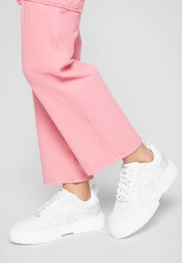 Vita sneakers med en tjock sula, slät läderöverkänsla och mesh-accenter. Bärs med rosa vida byxor, vilket framhäver skoens design.