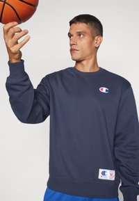 Sudadera navy con puños y dobladillo acanalados, que presenta un pequeño logo en el pecho y una etiqueta en la parte inferior. Sostenida junto a un baloncesto.