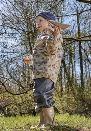 Bambino con giacca impermeabile chiara a motivi, polsini elastici, pantaloni impermeabili grigi e stivali di gomma beige, che tiene in mano un aeroplanino di carta.