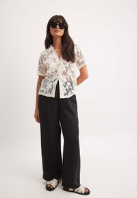 Witte kanten button-up shirt met korte mouwen, gecombineerd met zwarte wijde broek en zwart-witte sandalen. Model draagt oversized zwarte zonnebril.