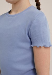 Svijetloplava rebrasta majica s okruglim izrezom i valovitim kratkim rukavima. Mekana tekstura, prilagođeni dizajn i glatka završna obrada tkanine.