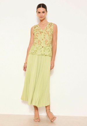 Femeie purtând un top fără mâneci din dantelă verde lime cu nasturi și o fustă midi plisată, stând cu mâinile relaxate pe un fundal alb.