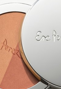 Ere Perez pudderblush med presset pulver i en metallisk sølvkompakt. Har en varm, teksturert ferskenfarge med preget logodetalj. Jevn finish.