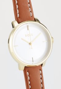 Reloj dorado con una esfera blanca, marcadores de hora minimalistas y correa de cuero marrón con detalles de costura blanca.