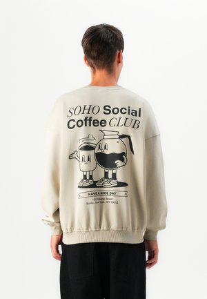 Person wearing beige sweatshirt with black cartoon coffee pot and cup characters and text “SOHO Social Coffee CLUB” on back.