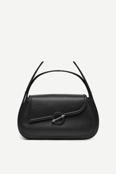 Sac à main en cuir noir avec rabat asymétrique et fermoir métallique circulaire, présentant deux fines poignées et un design épuré et minimaliste.