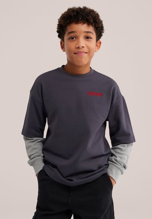 Jeune garçon aux cheveux bouclés porte un chemisier gris à manches longues en couches et un t-shirt sombre à manches courtes avec un texte rouge, debout les mains dans les poches.