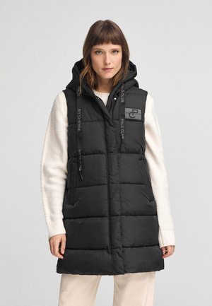 Zwarte puffer vest met een capuchon, voorzien van een gequiltde textuur, verticale naden, zijzakken en een logo op de borst; gecombineerd met een crèmekleurige trui.
