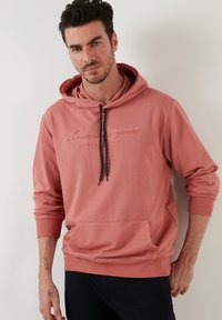 Rózsaszín kapucnis pulóver elülső zsebbel, húzózsinórral és fekete pettyes díszítéssel. A mellkason szöveges dizájn található, és testhezálló mandzsetták jellemzik. Puha anyagból készült.