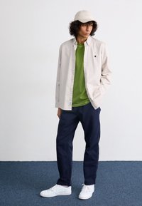 Cămașă bej cu nasturi, tricou verde, pantaloni bleumarin și adidași albi. Prezentă un logo pe cămașă și o șapcă de culoare deschisă.