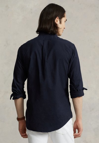 Uomo con capelli scuri di media lunghezza, che indossa una camicia blu scuro a maniche lunghe con le maniche arrotolate e pantaloni bianchi, visto da dietro.