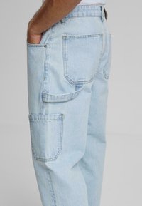 Pantaloni cargo din denim de culoare albastră deschisă, cu buzunare laterale și posterior, cu o croială lejeră și detalii de cusătură vizibile.