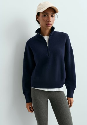 Vrouw die een marineblauwe trui met halve rits draagt, een wit overhemd eronder, grijze legging en een lichtbeige baseballpet tegen een effen witte achtergrond.