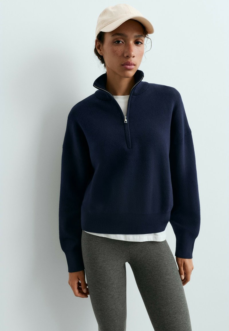 Femme portant un pull bleu marine à demi-fermeture éclair, une chemise blanche en dessous, des leggings gris et une casquette de baseball beige clair sur un fond blanc uni.