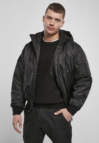 Giubbotto bomber nero con cappuccio staccabile, zip frontale e diverse tasche. Indossato sopra un maglione nero a trama e pantaloni scuri.