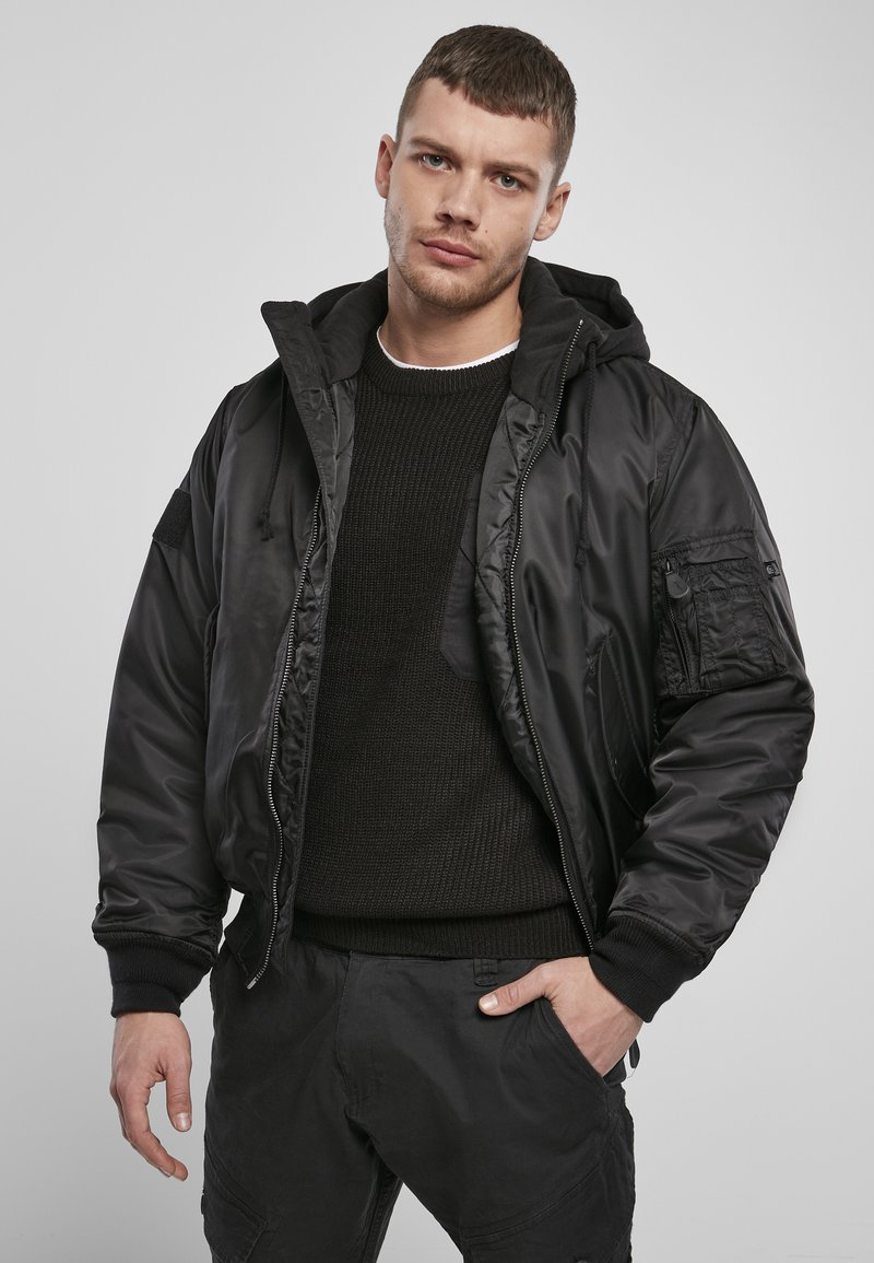 Giubbotto bomber nero con cappuccio staccabile, zip frontale e diverse tasche. Indossato sopra un maglione nero a trama e pantaloni scuri.