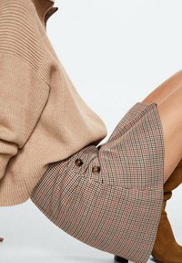 Personne portant un pull côtelé beige, une jupe à carreaux boutonnée et des bottes en daim marron, assise avec les jambes pliées sur fond blanc.