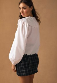 Fehér blúz nagy, hímzett gallérral és fodros ujjakkal, sötét tónusú, kockás szabott szoknyával párosítva.