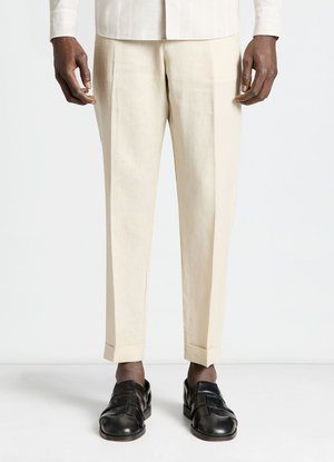 Homme portant un pantalon sur mesure couleur crème et des sandales en cuir noires à bouts fermés, debout devant un fond blanc uni.