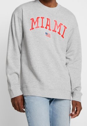Sweatshirt gris avec le texte "MIAMI" rouge et un petit graphique du drapeau américain. Il présente un col rond et des poignets et un ourlet côtelés.