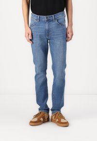 Jeans de mezclilla azul claro con un corte recto, diseño de cinco bolsillos y una sutil textura deslavada. Llevados con zapatillas color miel que tienen un upper texturizado.