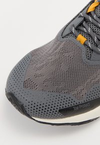 Baskets de sport grises avec une tige en mesh texturé, un capuchon en caoutchouc étiqueté "Protection des orteils" et des accents réfléchissants. Lacets en jaune contrasté.