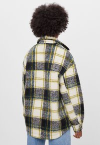 Rutigt skjorta i vit, gul, grå och grön flanell. Har en krage och långa ärmar med en avslappnad passform; synlig sömnad.